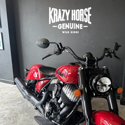 A red and black cruiser motorcycle photographed indoors against a dark gray wall with large white 'Krazy Horse Genuine Wild Rides' lettering. The bike is shown from a front three-quarter angle, with the motorcycle and branded wall visible.