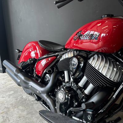 A close-up side view of a red Indian motorcycle, showing the glossy red fuel tank with the Indian script logo, black seat, black exhaust pipes, and silver engine components against a dark gray wall and concrete floor.