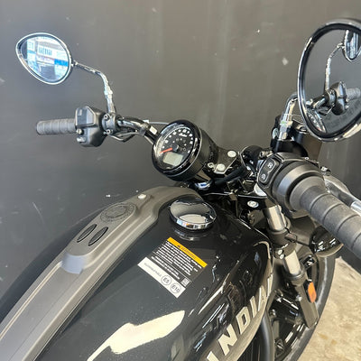 Close-up of a black Indian motorcycle showing the handlebars, round analog speedometer, fuel tank, and part of the front wheel against a dark gray indoor background.