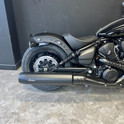 A close-up side view of a matte black Indian Scout motorcycle, focusing on the exhaust pipe, rear wheel, and seat against a dark gray wall and tiled floor.