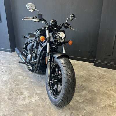 A black cruiser motorcycle photographed indoors on a light stone floor against a dark gray wall. The bike has chrome mirrors and forks, a round headlight, orange accent lights, and black wheels.