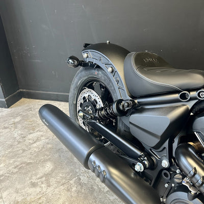 Close-up of a matte black cruiser motorcycle showing the rear wheel, fender, seat, exhaust pipe, and frame against a dark wall and concrete floor.