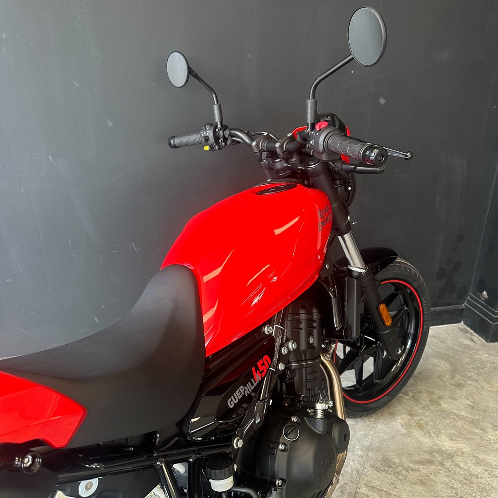 A red and black motorcycle photographed indoors against a dark gray wall and light concrete floor. The bike is shown from a side-rear angle, with the fuel tank, seat, mirrors, front wheel, and engine visible.