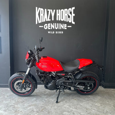 A red and black motorcycle parked indoors against a dark wall with the text 'Krazy Horse Genuine Wild Rides' above it.