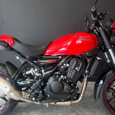 A Royal Enfield Bullet S motorcycle with a vibrant red fuel tank and black body components, photographed from a side angle against a dark gray wall.