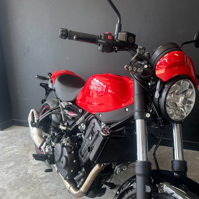 A red and black Triumph motorcycle photographed indoors against a dark gray wall and light gray floor. The bike is shown at a three-quarter front angle, with the handlebars, round headlight, fuel tank, engine, exhaust, and front fork visible.