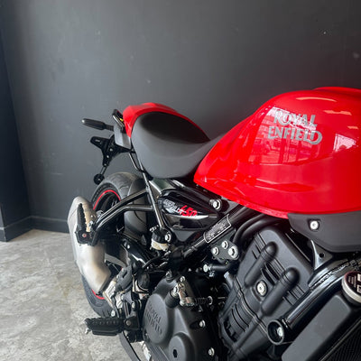 A close-up side view of a red and black motorcycle, showing the fuel tank, seat, exhaust, and engine against a dark gray wall and concrete floor.