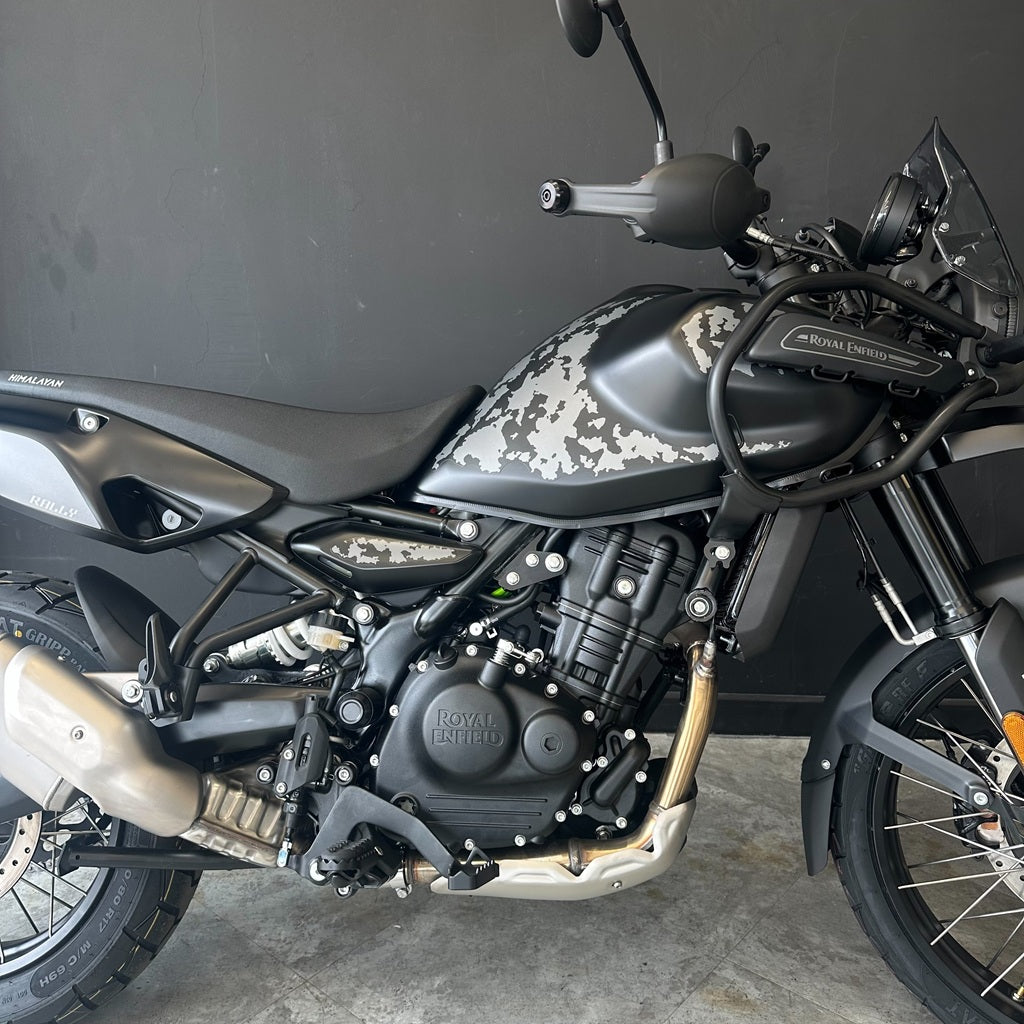 A Royal Enfield motorcycle with a matte black and gray camouflage pattern on the fuel tank and side panels, photographed indoors against a dark gray wall.