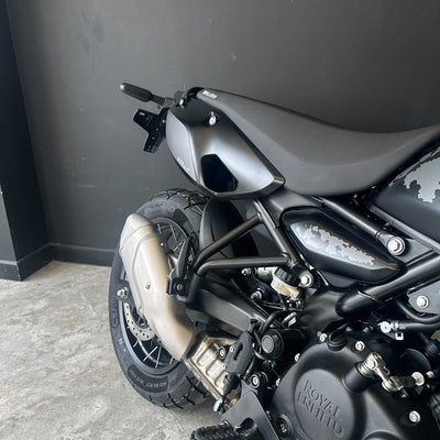 Close-up rear three-quarter view of a matte black Royal Enfield motorcycle, showing the rear fender, exhaust, wheel, and engine against a dark gray wall and concrete floor.