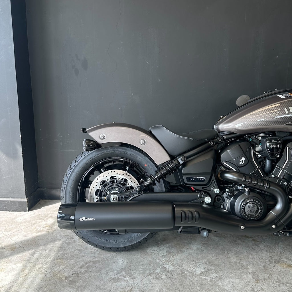 A side view of a cruiser motorcycle, showing the rear wheel, black exhaust muffler with a visible 'Matich' logo, black seat, and metallic brown/bronze fuel tank against a dark gray wall.