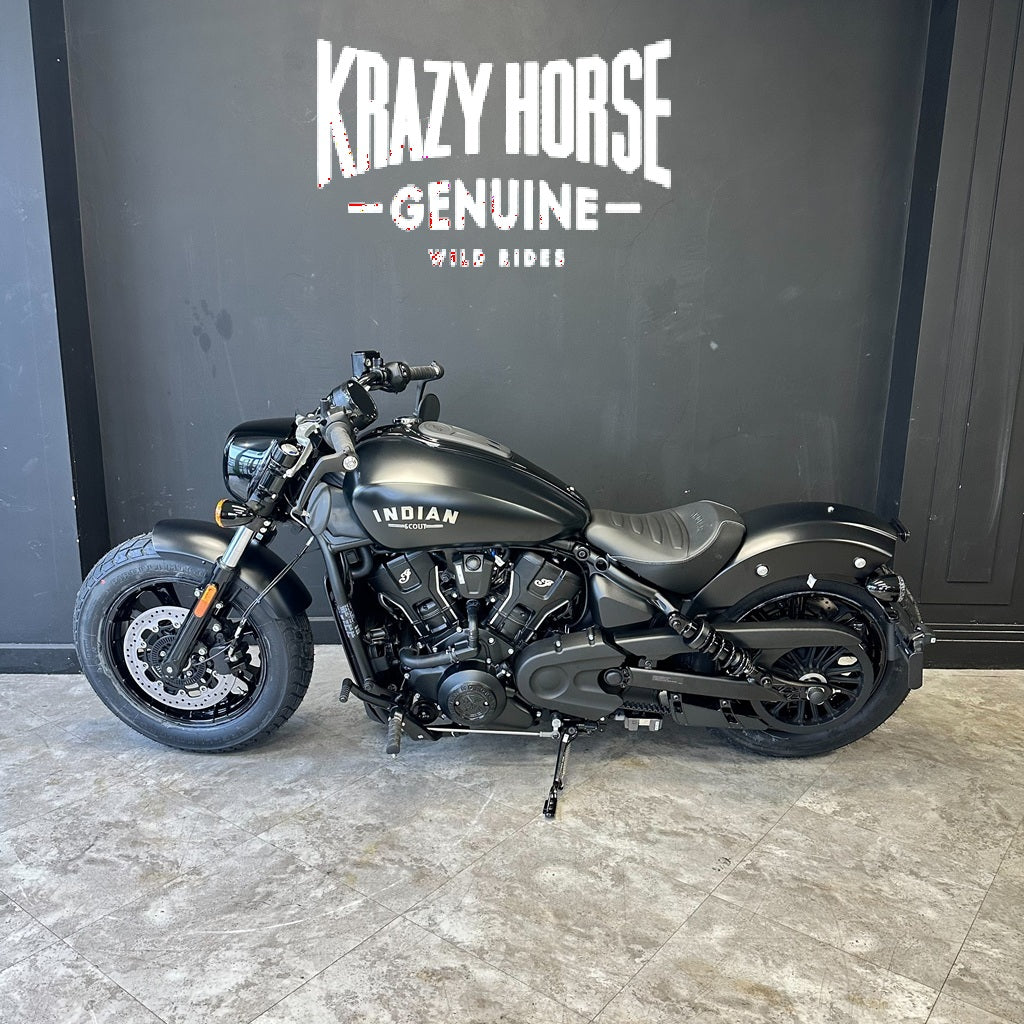 A matte black Indian motorcycle with a cruiser-style design, parked indoors against a dark gray wall with 'Krazy Horse Genuine Wild Ride' branding. The bike has a black seat, black wheels, and visible engine components.