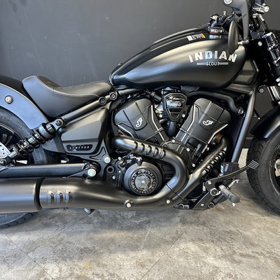 A black Indian Scout motorcycle photographed indoors against a dark gray wall and gray tiled floor, showing the side fuel tank, engine, exhaust, seat, and part of the front wheel.