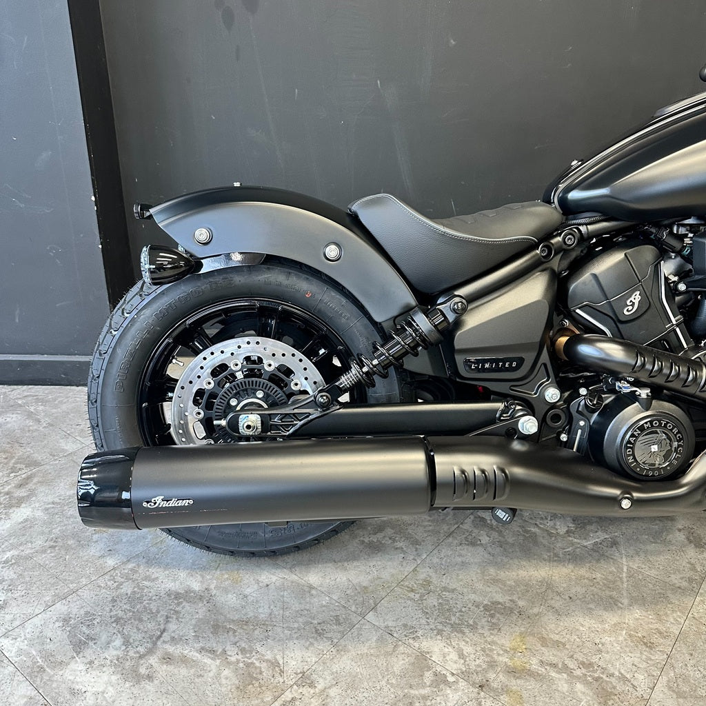 A matte black Indian Scout motorcycle shown from the rear three-quarter view, featuring a matching black exhaust muffler with the Indian logo, set against a dark gray wall and gray tiled floor.