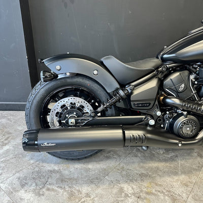 A matte black Indian Scout motorcycle shown from the rear three-quarter view, featuring a matching black exhaust muffler with the Indian logo, set against a dark gray wall and gray tiled floor.