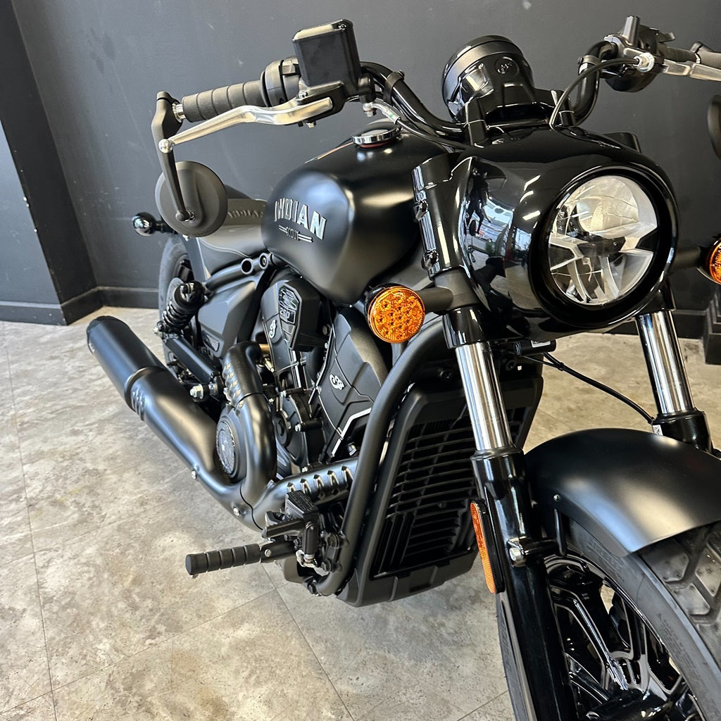 A black Indian motorcycle photographed indoors on a gray tiled floor against a dark gray wall. The bike is shown at a front three-quarter angle, with the Indian logo visible on the fuel tank.