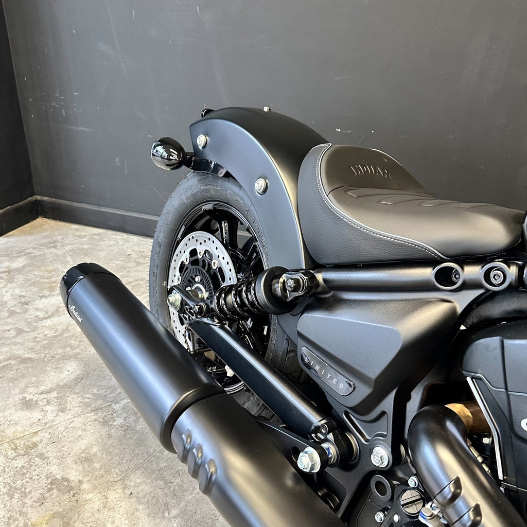 Close-up of a matte black cruiser motorcycle showing the rear fender, black seat with embossed branding, rear wheel, disc brake, and dual exhaust pipes against a dark gray wall and concrete floor.