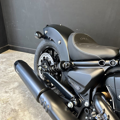 Close-up of a matte black cruiser motorcycle showing the rear fender, black seat with embossed branding, rear wheel, disc brake, and dual exhaust pipes against a dark gray wall and concrete floor.