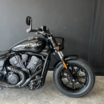 A black Indian Scout motorcycle photographed indoors against a dark gray wall and concrete floor, showing the side profile with tank, engine, wheel, and handlebars visible.