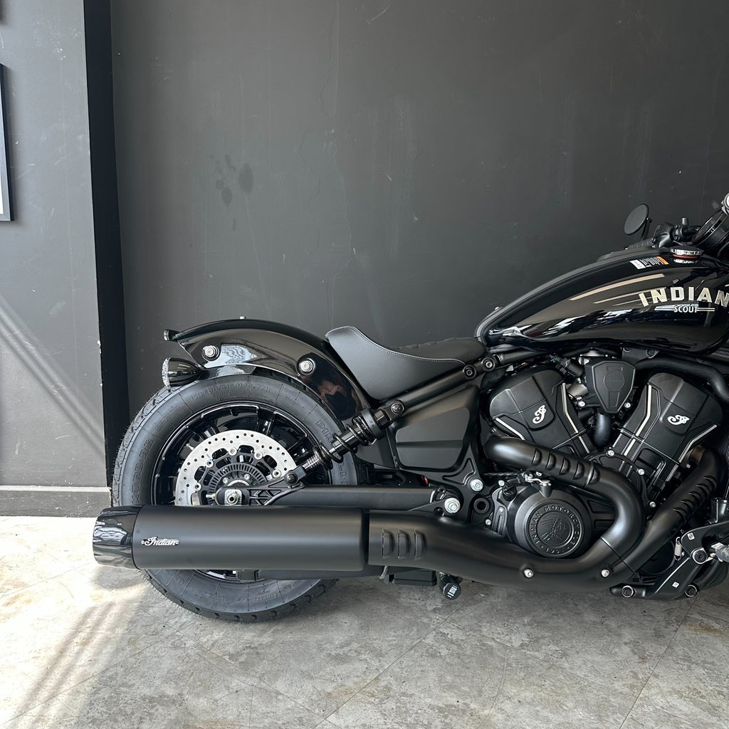 A black Indian Chopper motorcycle photographed indoors against a dark gray wall, showing the side profile with the exhaust, wheel, engine, and part of the tank visible.