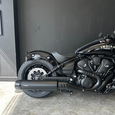 A black Indian Chopper motorcycle photographed indoors against a dark gray wall, showing the side profile with the exhaust, wheel, engine, and part of the tank visible.