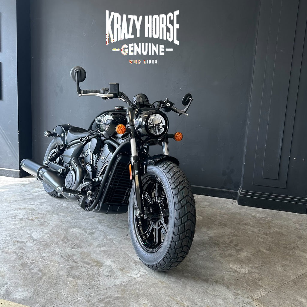 A black Harley-Davidson cruiser motorcycle is parked indoors against a dark gray wall with the text 'Krazy Horse Genuine Rider' visible above it. The bike is shown at a three-quarter front angle on a concrete floor.