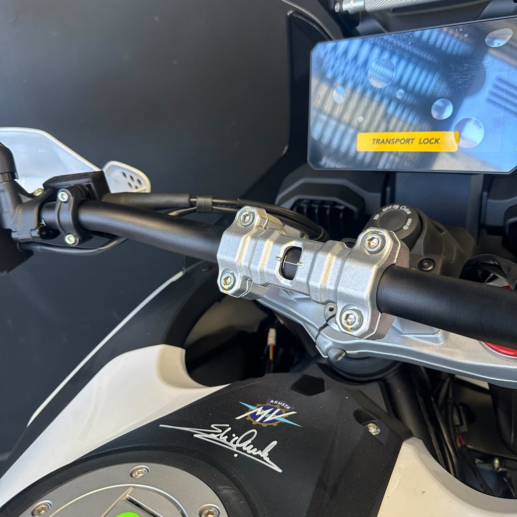 Close-up of a motorcycle handlebar area with a silver clamp mounted on black handlebars, a white and black fuel tank with a visible stylized logo, and part of a blue digital display screen.