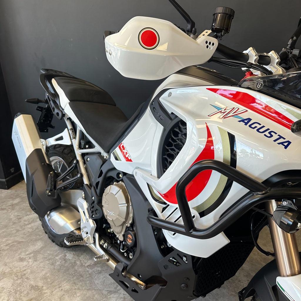 A white Agusta motorcycle with red, black, and gold graphics, featuring a black seat, black crash bar, and a white hardtop storage box, photographed in a garage setting.