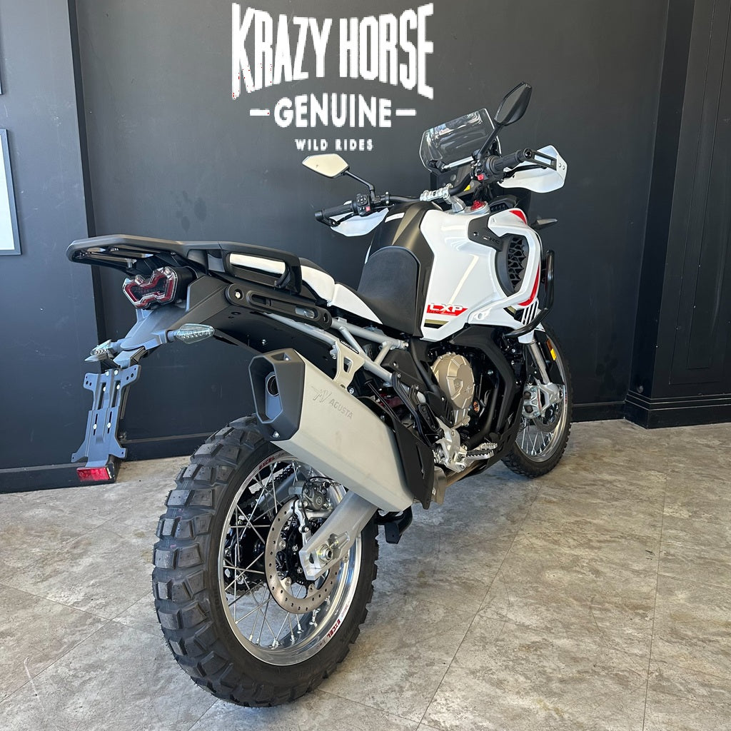 A white and black adventure motorcycle with red accents, photographed indoors against a dark wall with 'Krazy Horse Genuine Wild Ride' signage. The bike is shown from a rear three-quarter angle, highlighting the tail light, exhaust, rear suspension, and side profile.