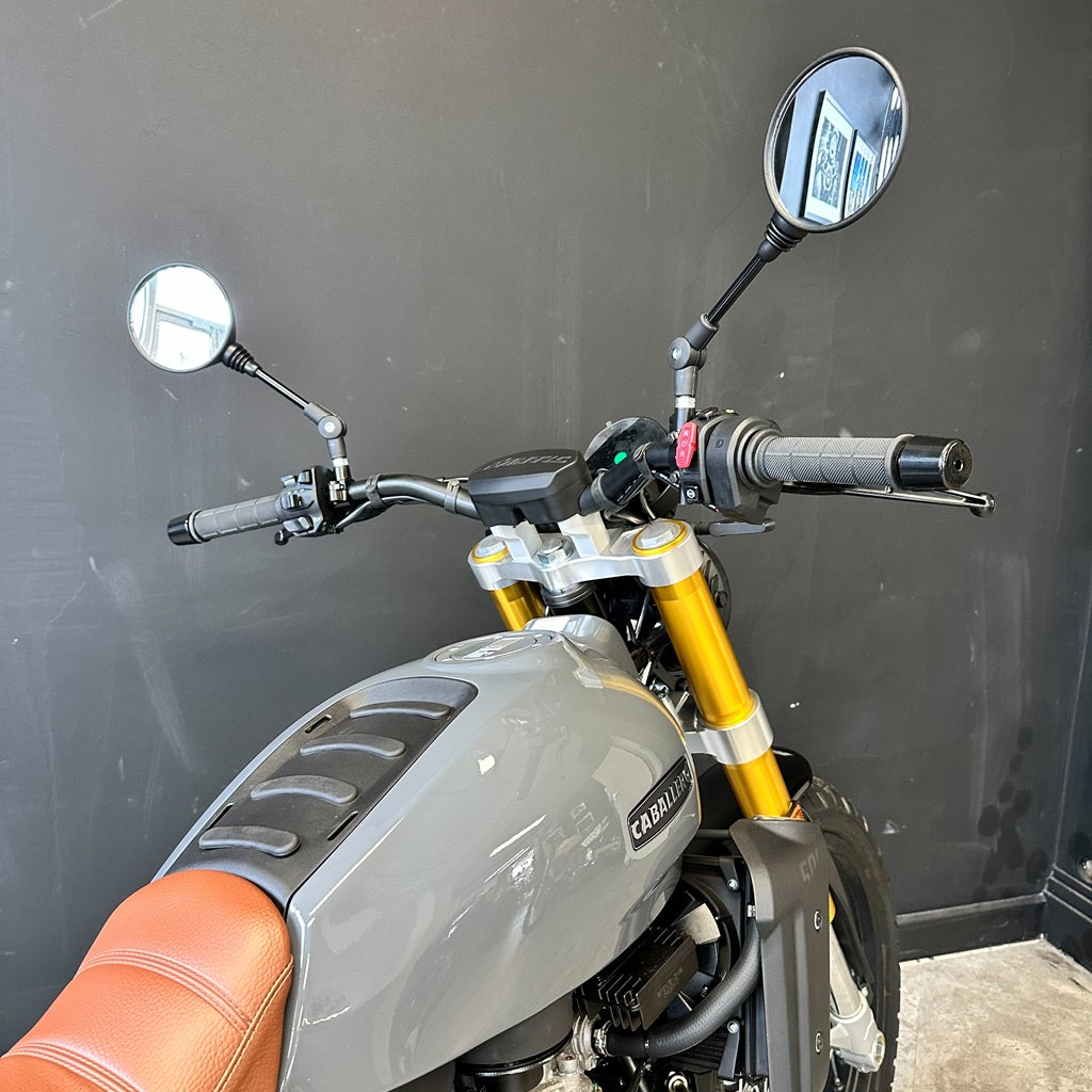 Close-up of a motorcycle's handlebar area, showing a gray fuel tank with a visible 'Cagla' brand badge, bright gold front forks, black grips, and two round mirrors against a dark gray wall.