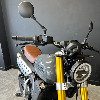 A close-up of a motorcycle front section, showing a gray fuel tank with 'MUMBAO' branding, a brown seat, gold front forks, black handlebars, and mirrors, photographed indoors against a dark gray wall.