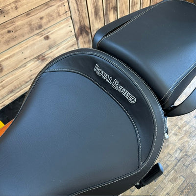 Close-up of a black motorcycle seat with white stitching and the embossed brand name 'ROYAL ENFIELD' on the backrest, set against a rustic wooden background.