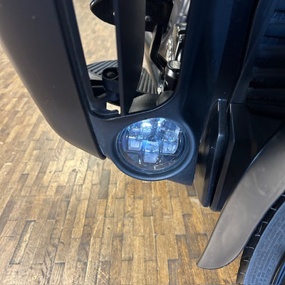 A close-up of a motorcycle's front headlight assembly, showing the light turned on with a bright blue-white glow, mounted on a black fender over a wood plank floor.