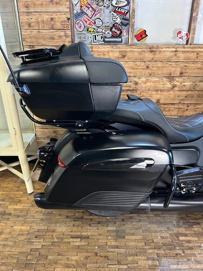 A black Harley-Davidson touring motorcycle with a large hardtop storage box and dual saddle, photographed indoors against a wood-paneled wall with stickers and a brick floor.
