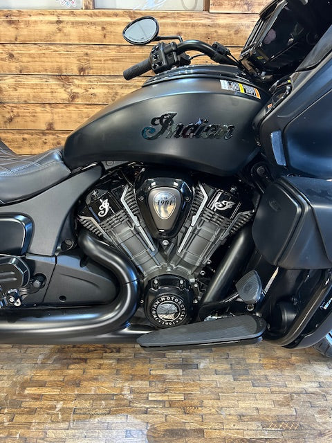 A close-up side view of a black Indian motorcycle, showing the fuel tank with the 'Indian' script logo, V-twin engine, and exhaust pipes, set against a wooden wall and floor.