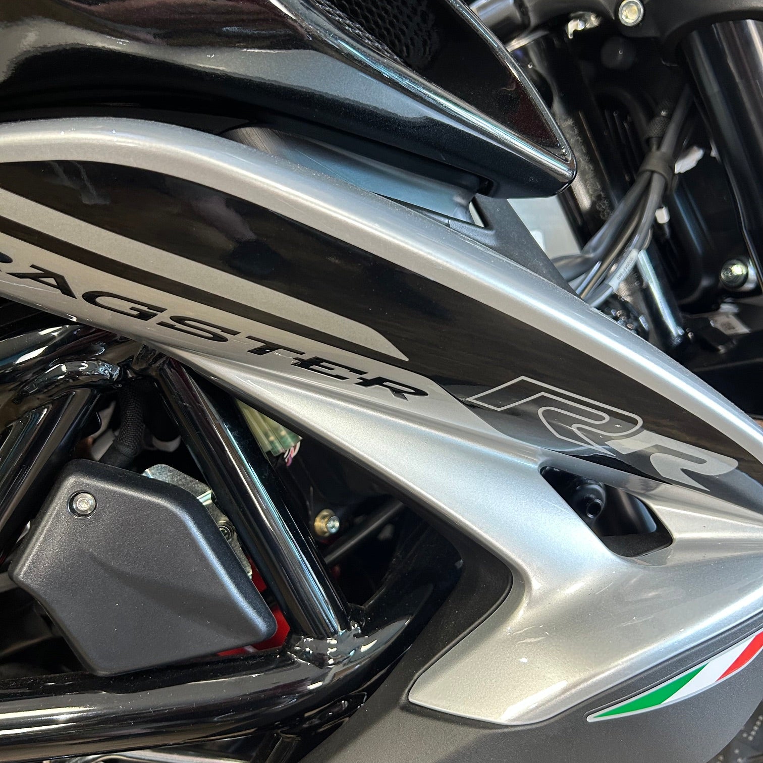 Close-up of a motorcycle body panel with the word 'AGSTER' and 'R2' visible, showing a metallic silver and black finish with a small green, white, and red accent near the lower right.