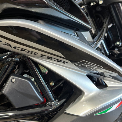 Close-up of a motorcycle body panel with the word 'AGSTER' and 'R2' visible, showing a metallic silver and black finish with a small green, white, and red accent near the lower right.
