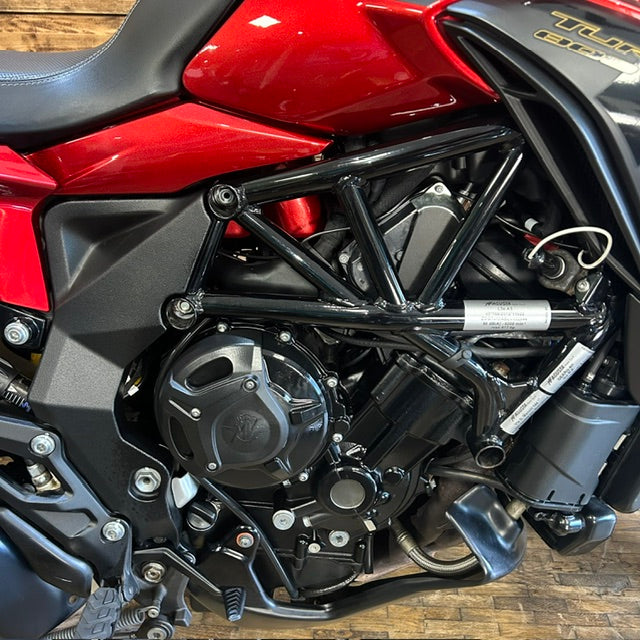 Close-up of a red and black motorcycle engine and frame, with visible mechanical components and a wood floor in the background.