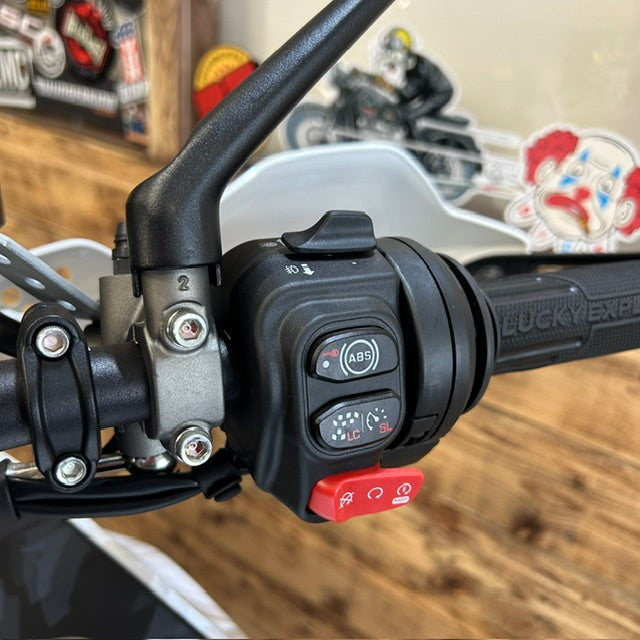 Close-up of a motorcycle handlebar with black controls, a red switch, and a silver brake lever on a wooden table with stickers in the background.