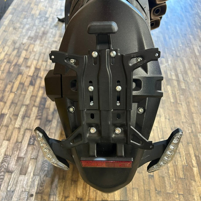 A black metal motorcycle rear rack with mounting brackets and silver screws, attached to the rear of a black motorcycle, photographed on a wooden floor.