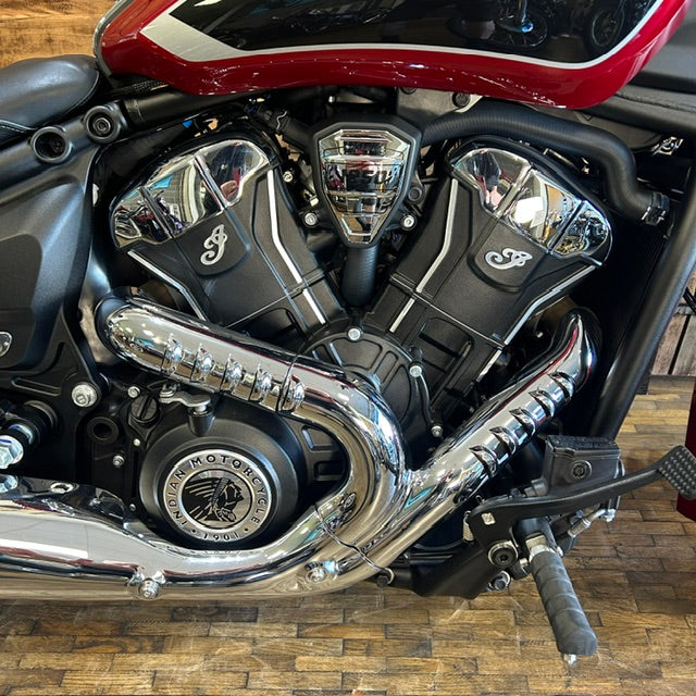Close-up of a motorcycle engine with polished chrome exhaust pipes, black V-tube covers, and a red and black fuel tank on a wooden floor.