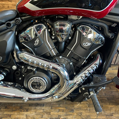 Close-up of a motorcycle engine with polished chrome exhaust pipes, black V-tube covers, and a red and black fuel tank on a wooden floor.